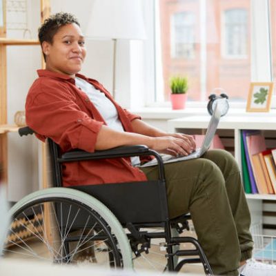 Portrait of African young woman with disabilities sitting in wheelchair and looking at camera while using laptop computer at home
